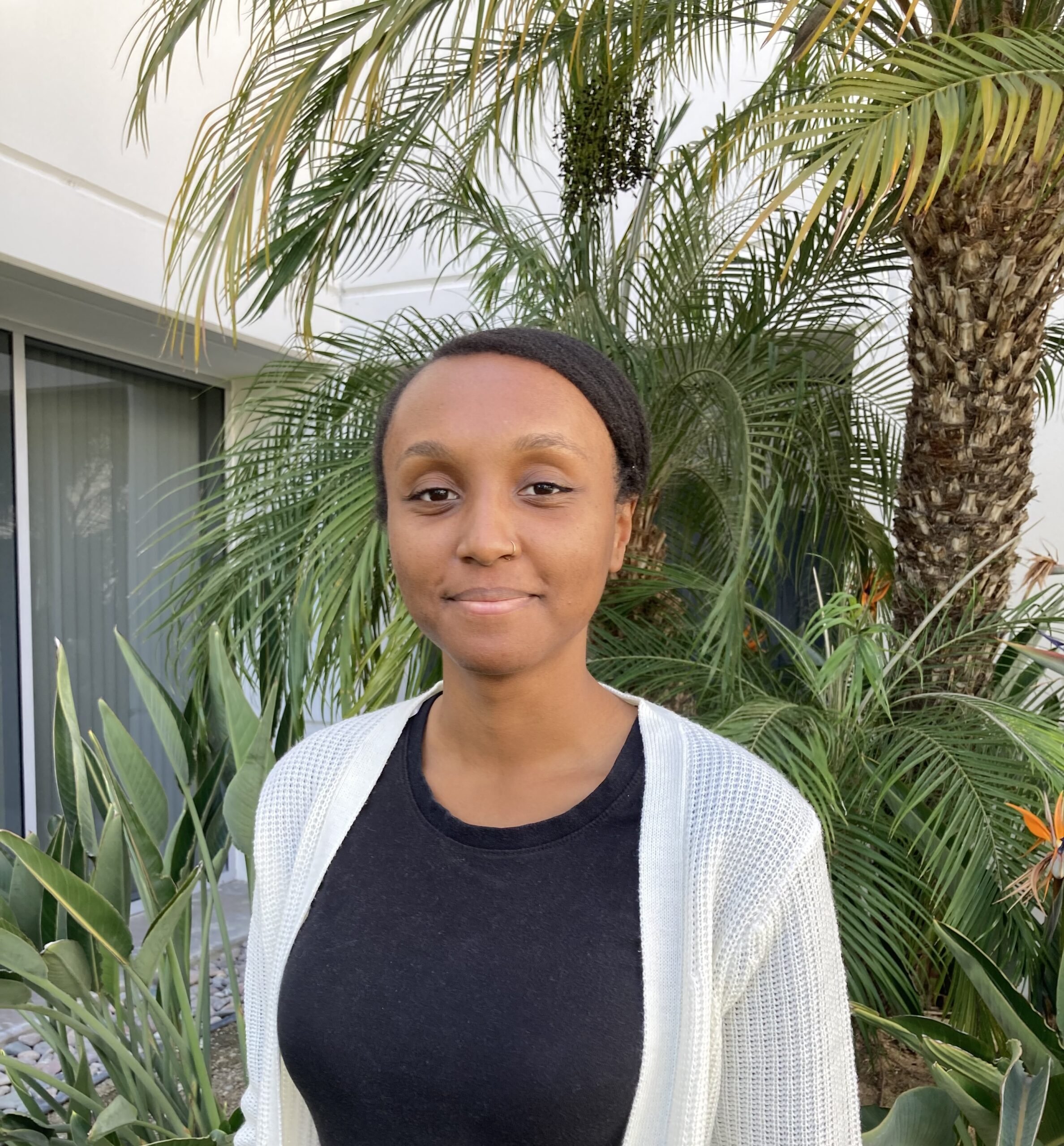 African American, Black female standing in front of green plant in front of white building.  She has on a white cotton sweater over a black shirt.